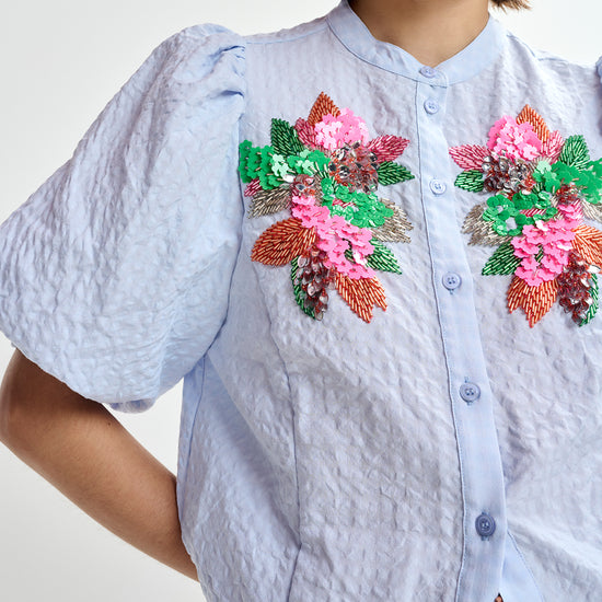 Woman wearing a light blue embroidered shirt with floral designs by Essentiel Antwerp and brown pants on a white background close up