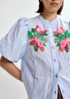 Woman wearing a light blue embroidered shirt with floral designs by Essentiel Antwerp and brown pants on a white background close up