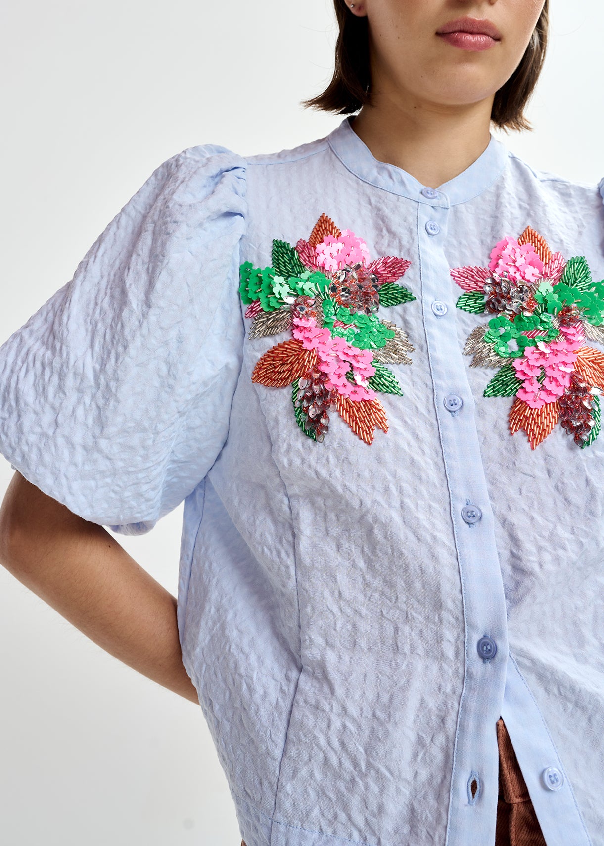 Woman wearing a light blue embroidered shirt with floral designs by Essentiel Antwerp and brown pants on a white background close up