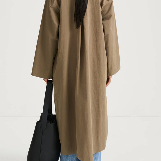 A model shown from behind wearing a long, oversized taupe-brown coat over light-wash blue jeans, holding a black tote bag against a plain white studio background.