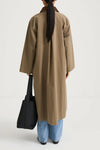 A model shown from behind wearing a long, oversized taupe-brown coat over light-wash blue jeans, holding a black tote bag against a plain white studio background.