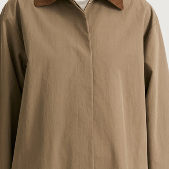 A close-up of a person wearing an oversized taupe-brown button-up shirt jacket with a contrasting brown suede-like collar, photographed against a light gray studio background.