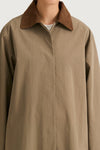 A close-up of a person wearing an oversized taupe-brown button-up shirt jacket with a contrasting brown suede-like collar, photographed against a light gray studio background.