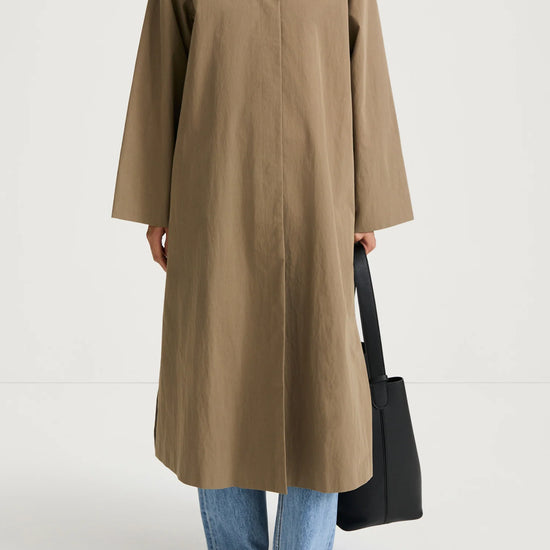 A model wearing a long, oversized taupe-brown coat with a contrasting dark brown collar, styled with light-wash blue jeans, black flats, and a black tote bag against a plain white studio background.