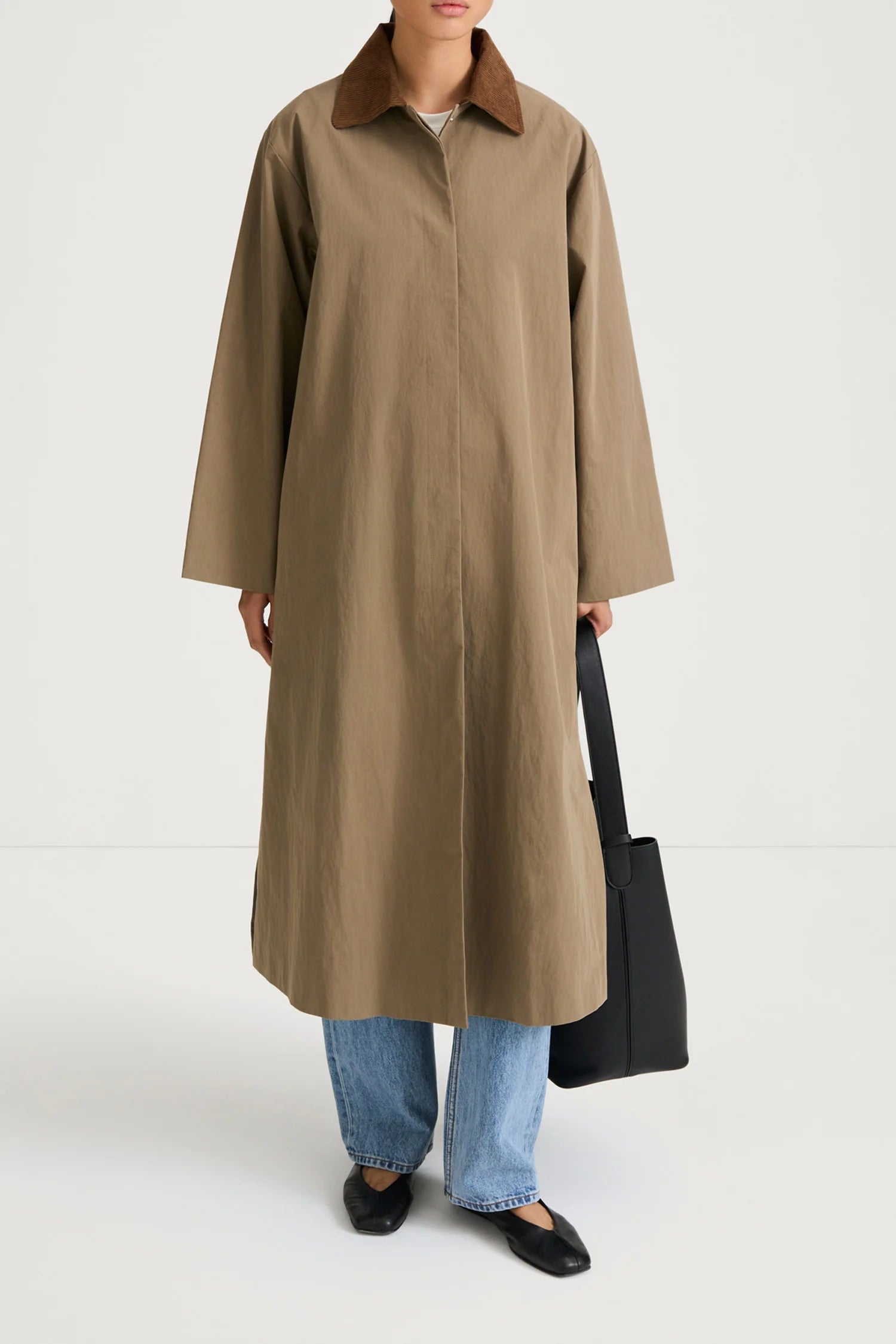 A model wearing a long, oversized taupe-brown coat with a contrasting dark brown collar, styled with light-wash blue jeans, black flats, and a black tote bag against a plain white studio background.