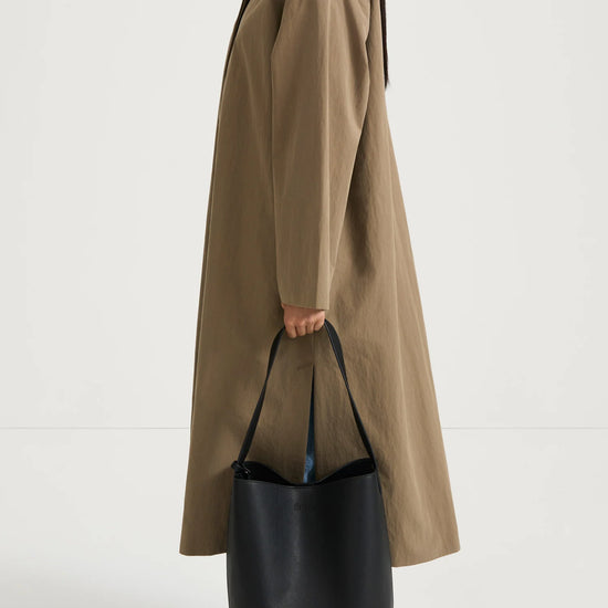 A model shown from the side wears a long, oversized taupe-brown coat with a contrasting dark brown collar, holding a large black leather bucket bag. The model is also wearing light-wash blue jeans and black flat shoes, set against a plain white studio background.