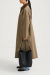 A model shown from the side wears a long, oversized taupe-brown coat with a contrasting dark brown collar, holding a large black leather bucket bag. The model is also wearing light-wash blue jeans and black flat shoes, set against a plain white studio background.