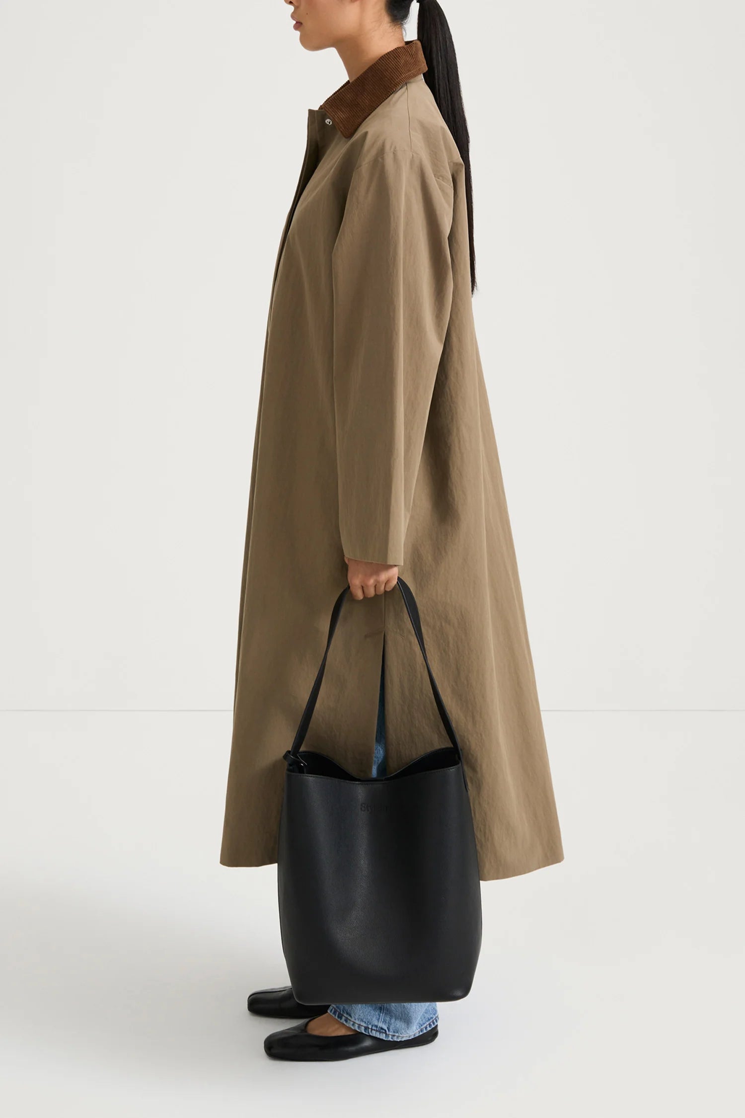 A model shown from the side wears a long, oversized taupe-brown coat with a contrasting dark brown collar, holding a large black leather bucket bag. The model is also wearing light-wash blue jeans and black flat shoes, set against a plain white studio background.