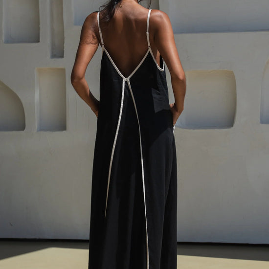 Reverse of Woman wearing a black dress with white straps against a light-coloured wall.