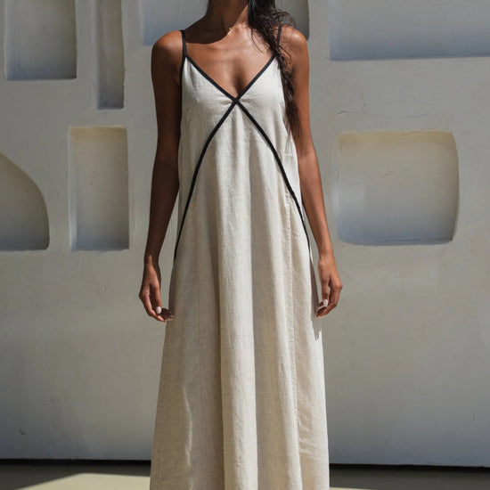 Woman wearing an oatmeal linen dress with black straps against a light-colored wall.