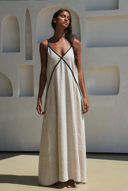 Woman wearing an oatmeal linen dress with black straps against a light-colored wall.