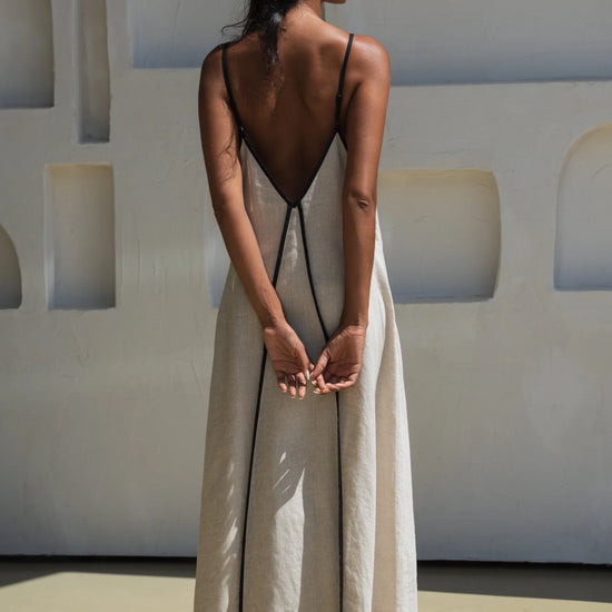 Woman wearing a long, light-colored linen dress with thin straps against a white wall.