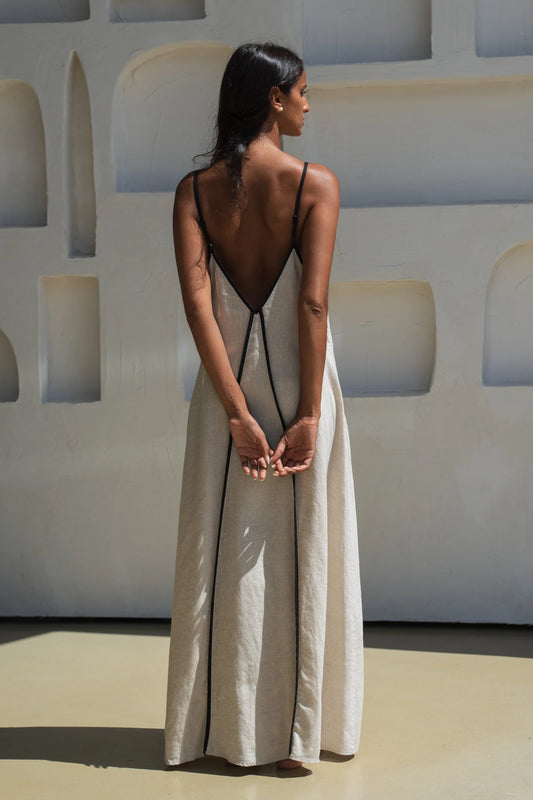 Woman wearing a long, light-colored linen dress with thin straps against a white wall.
