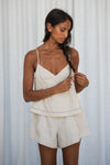 Woman wearing a beige linen top and shorts set against a white curtain background