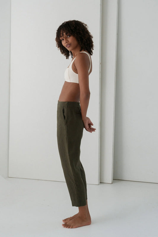 Woman wearing a white top and green linen trousers standing against a white wall.
