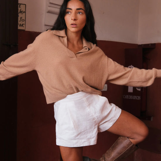 Woman in a camel sweater and white shorts posing in a room with a red wall.