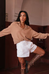 Woman in a camel sweater and white shorts posing in a room with a red wall.