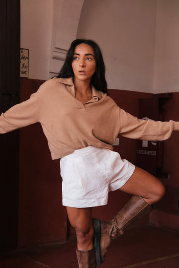 Woman in a camel sweater and white shorts posing in a room with a red wall.