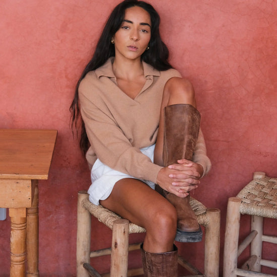 Woman sitting on a wooden stool against a pink wall wearing a camel shirt and brown knee-high boots.