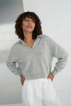 Woman wearing a gray sweater and white pants against a neutral background