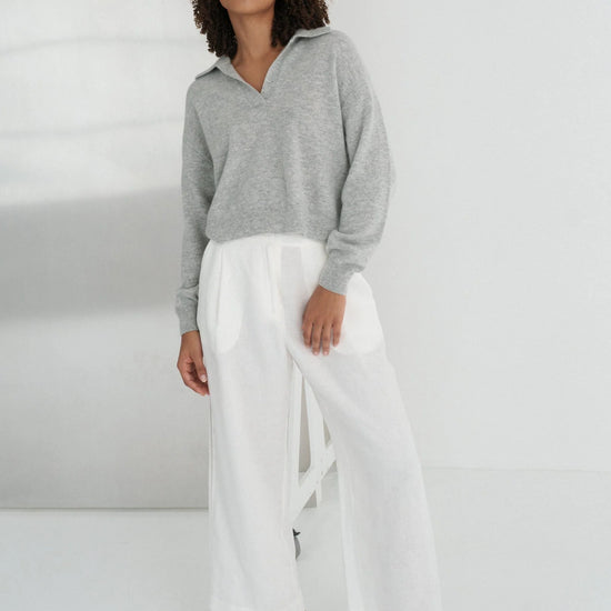Person wearing a gray sweater and white pants standing against a white wall with a reflective surface.