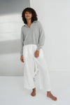 Person wearing a gray sweater and white pants standing against a white wall with a reflective surface.