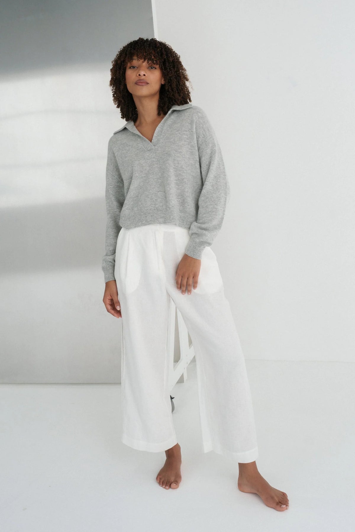 Person wearing a gray sweater and white pants standing against a white wall with a reflective surface.