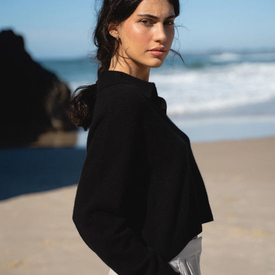 Woman in black jacket and white pants standing on a beach with ocean in the background