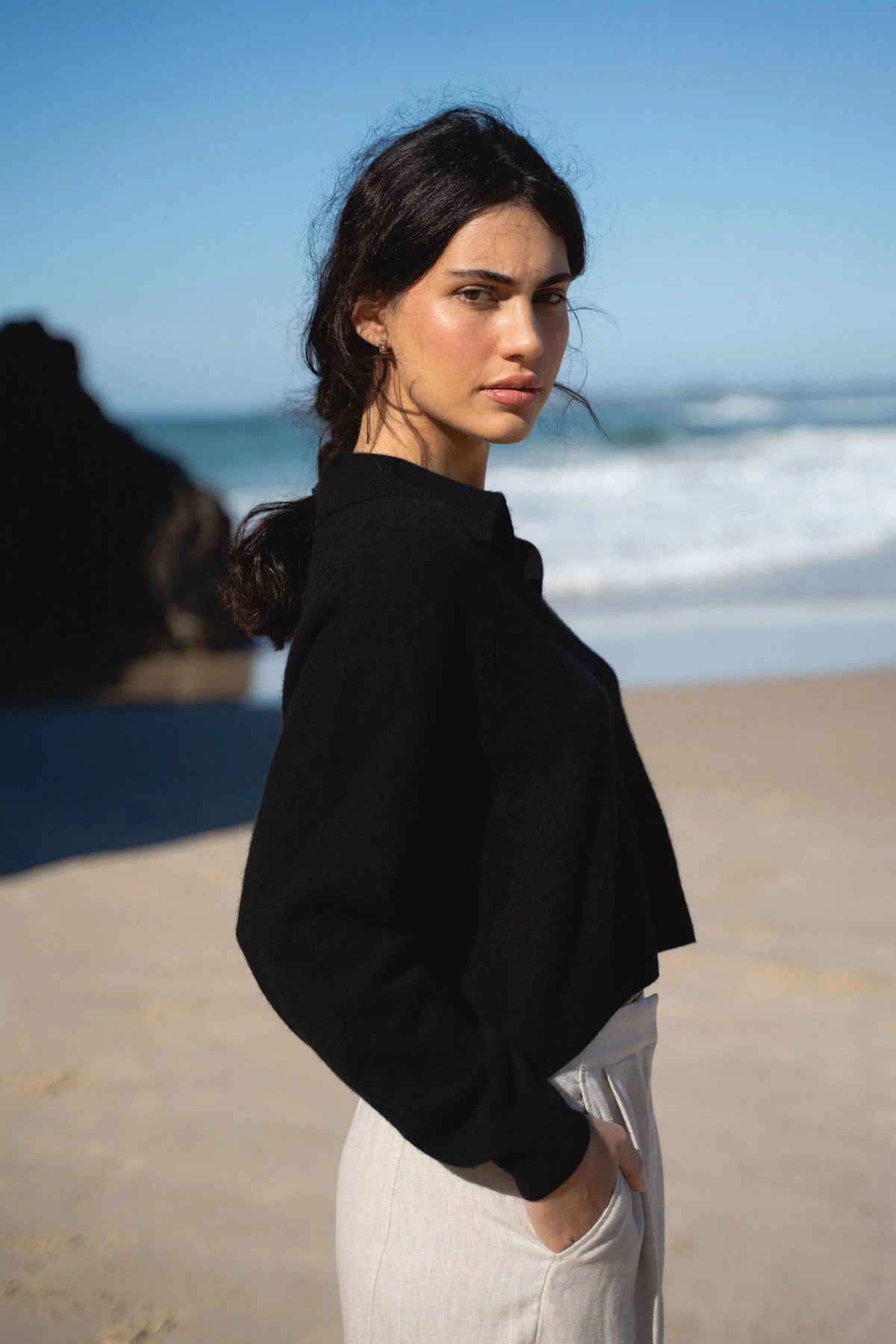 Woman in black jacket and white pants standing on a beach with ocean in the background