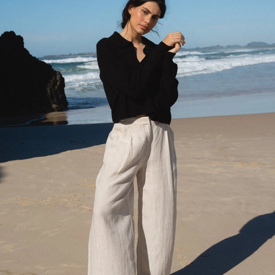 Woman in black sweater and light pants standing on a beach with ocean waves in the background