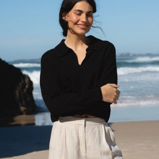Woman standing on a beach wearing a black top and light-colored pants with ocean waves in the background.