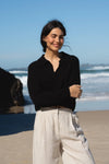 Woman standing on a beach wearing a black top and light-colored pants with ocean waves in the background.