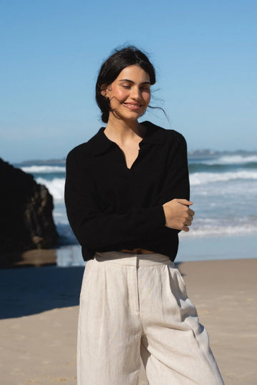 Woman standing on a beach wearing a black top and light-colored pants with ocean waves in the background.