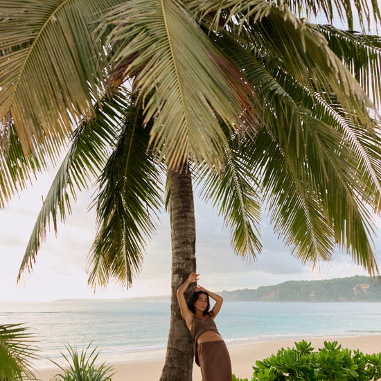 A woman in a brown two-piece outfit poses leaning against a large palm tree on a sandy beach with the ocean and distant land visible in the background.