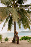 A woman in a brown two-piece outfit poses leaning against a large palm tree on a sandy beach with the ocean and distant land visible in the background.