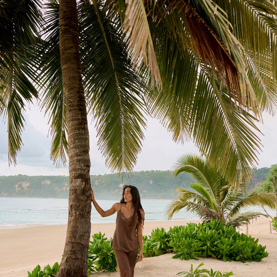 A woman stands on a sandy beach under a large palm tree, wearing a matching brown lounge set. The background features other palm trees, green shrubs, and the ocean under a cloudy sky.