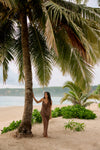 A woman stands on a sandy beach under a large palm tree, wearing a matching brown lounge set. The background features other palm trees, green shrubs, and the ocean under a cloudy sky.