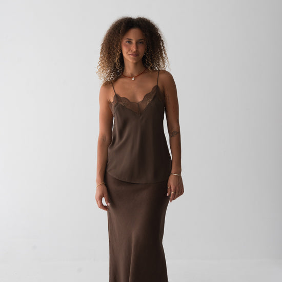 A woman stands barefoot against a plain light gray studio background wearing a matching two-piece lounge set in dark brown. The set includes a camisole top with lace trim at the neckline and a long skirt.