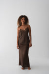 A woman stands barefoot against a plain light gray studio background wearing a matching two-piece lounge set in dark brown. The set includes a camisole top with lace trim at the neckline and a long skirt.