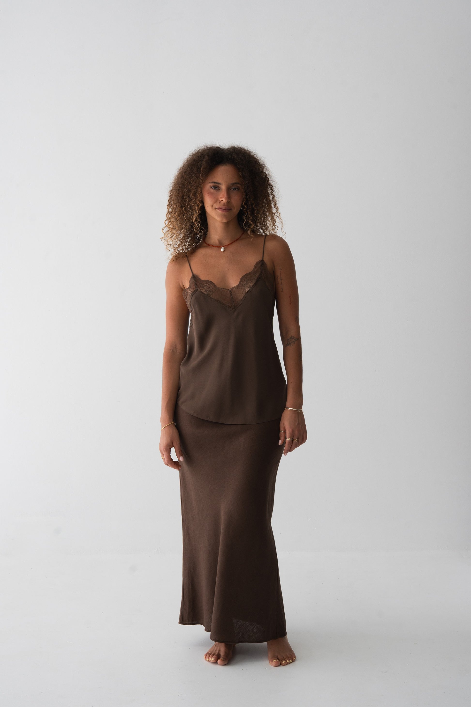 A woman stands barefoot against a plain light gray studio background wearing a matching two-piece lounge set in dark brown. The set includes a camisole top with lace trim at the neckline and a long skirt.