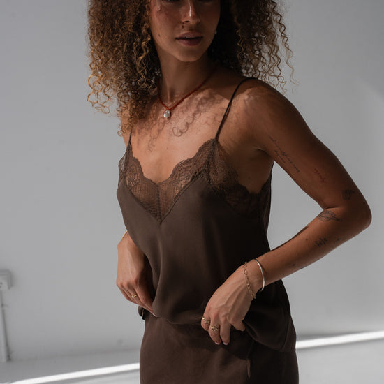 A woman with curly brown hair wearing a dark brown satin slip dress with a lace V-neckline, posing in a studio setting.