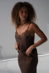 A woman with curly brown hair wearing a dark brown satin slip dress with a lace V-neckline, posing in a studio setting.