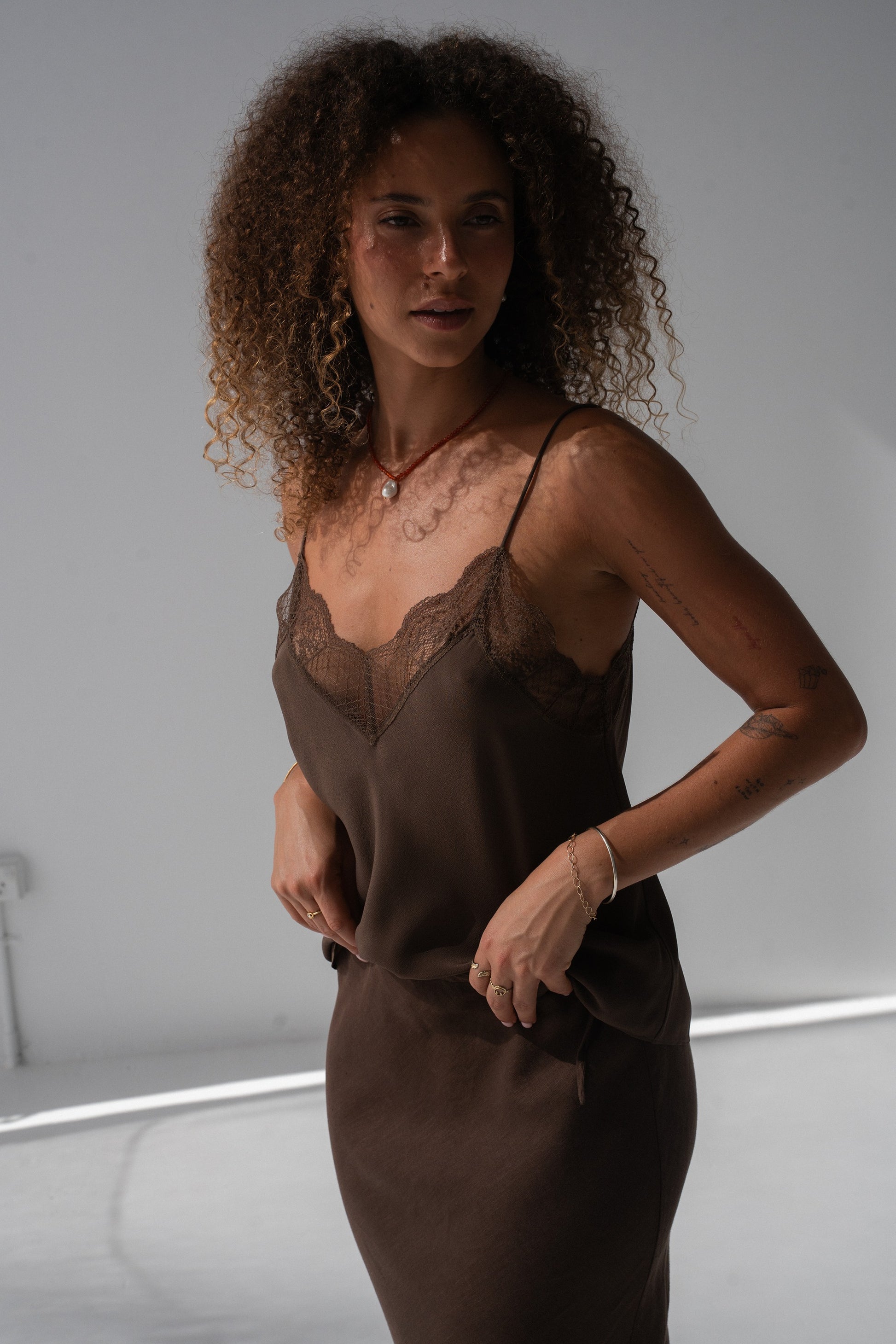 A woman with curly brown hair wearing a dark brown satin slip dress with a lace V-neckline, posing in a studio setting.