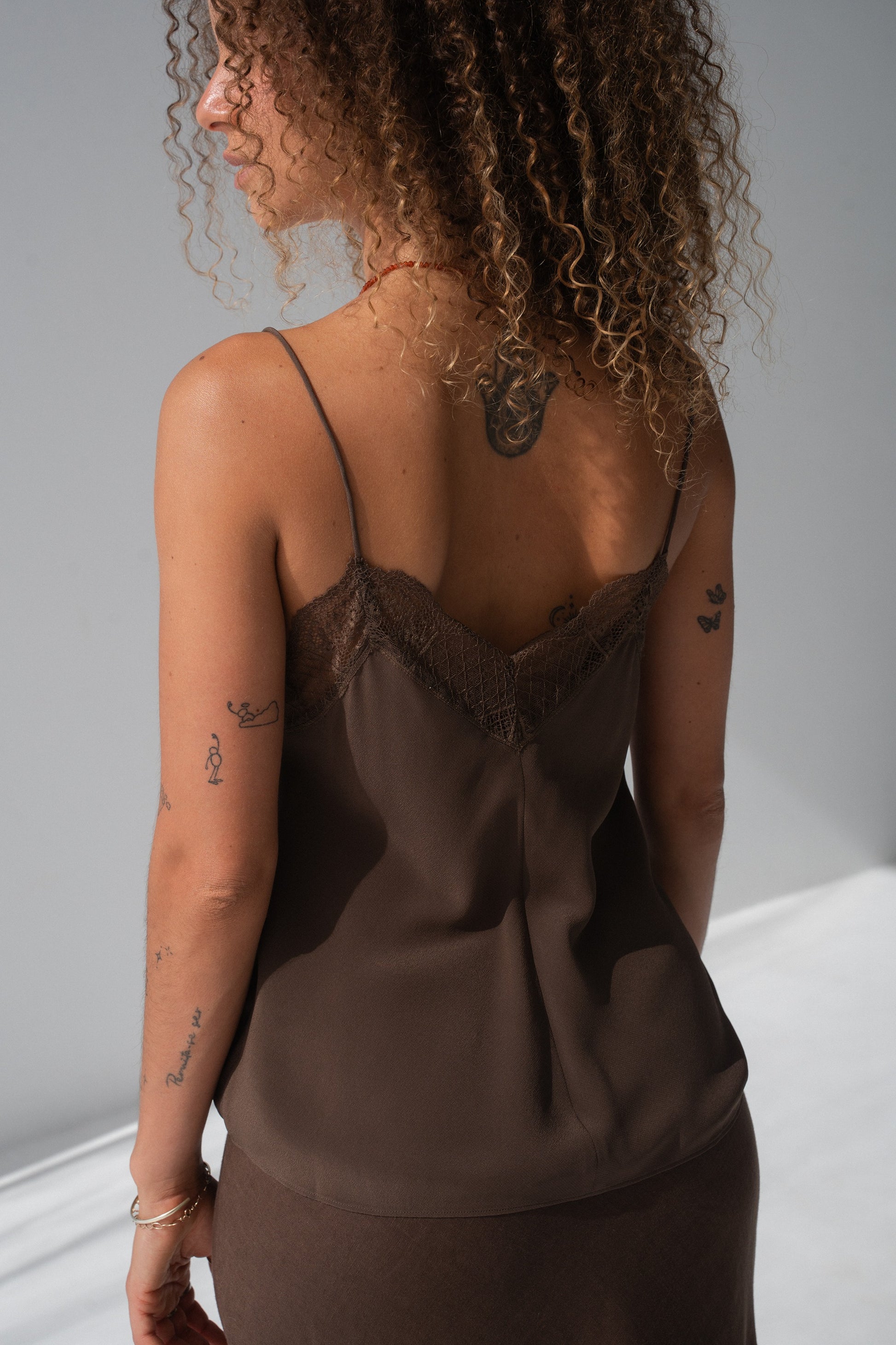 A woman shown from the back wearing a brown satin camisole with lace trim at the neckline, photographed in a studio setting.