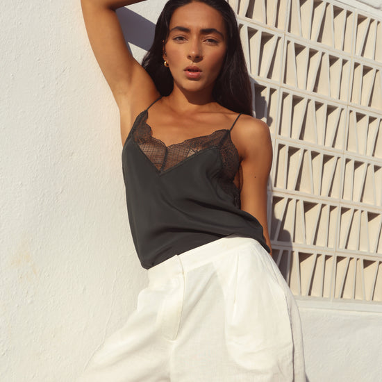 A woman posing in a black lace-trimmed camisole top and white wide-leg trousers against a textured white wall with a geometric window.