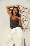 A woman posing in a black lace-trimmed camisole top and white wide-leg trousers against a textured white wall with a geometric window.