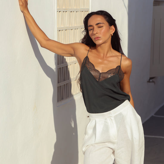 A woman posing outdoors against a white wall, wearing a dark charcoal gray camisole top with a sheer black lace bust and white wide-leg pants.