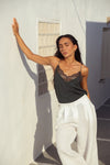 A woman posing outdoors against a white wall, wearing a dark charcoal gray camisole top with a sheer black lace bust and white wide-leg pants.