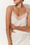 A close-up of a model wearing a white camisole top with thin straps, a lace V-neckline, and a matching white skirt against a clean white background.