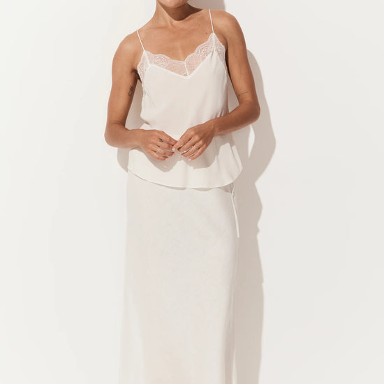 A woman modeling a two-piece white lingerie set consisting of a camisole top with lace trim and a matching long skirt, set against a plain white studio background.